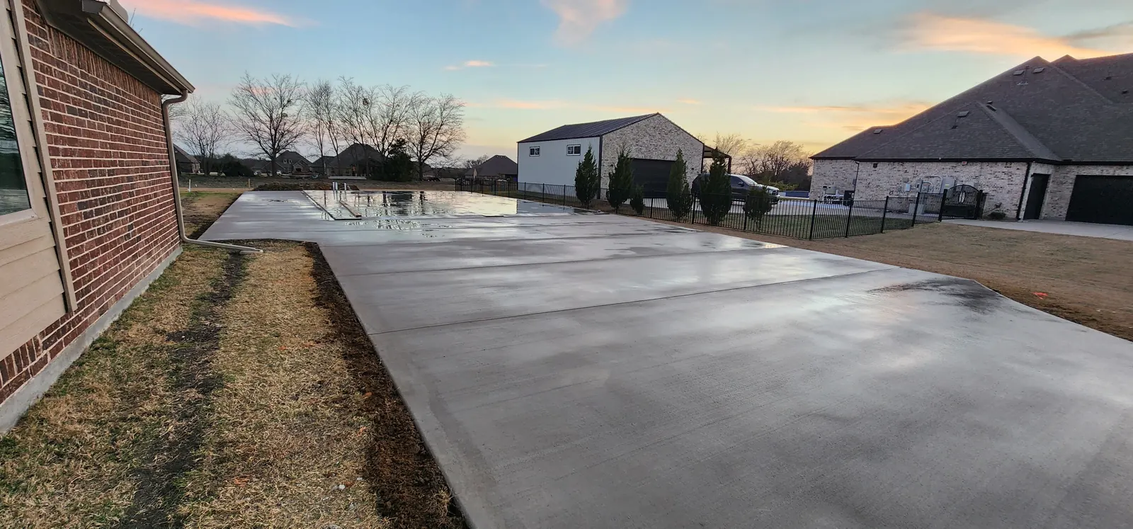 New concrete shop slab and driveway at sunset, with a metal shop and brick home in the background