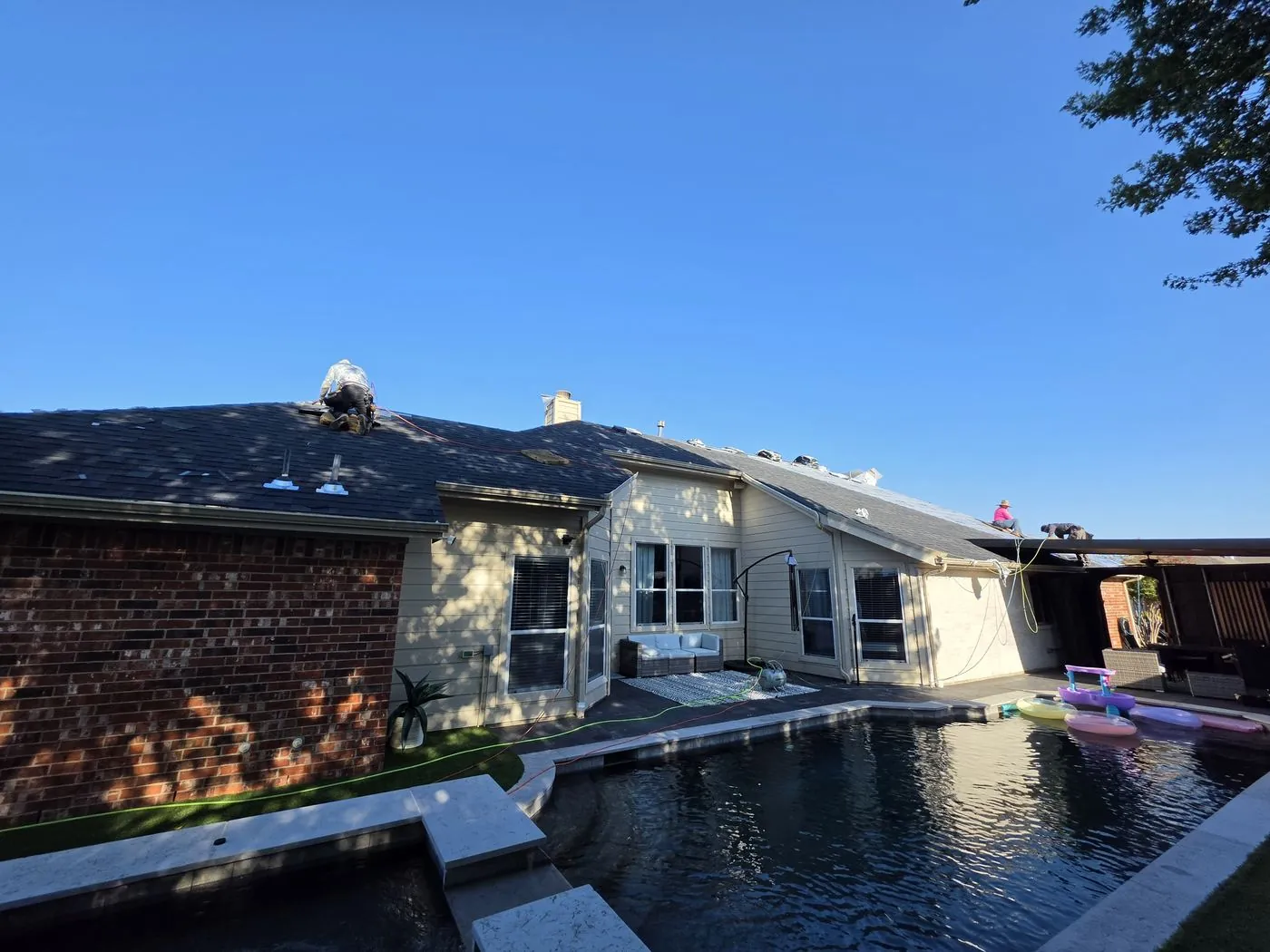 Roofing crew replacing shingles on a large single-story home overlooking a backyard pool