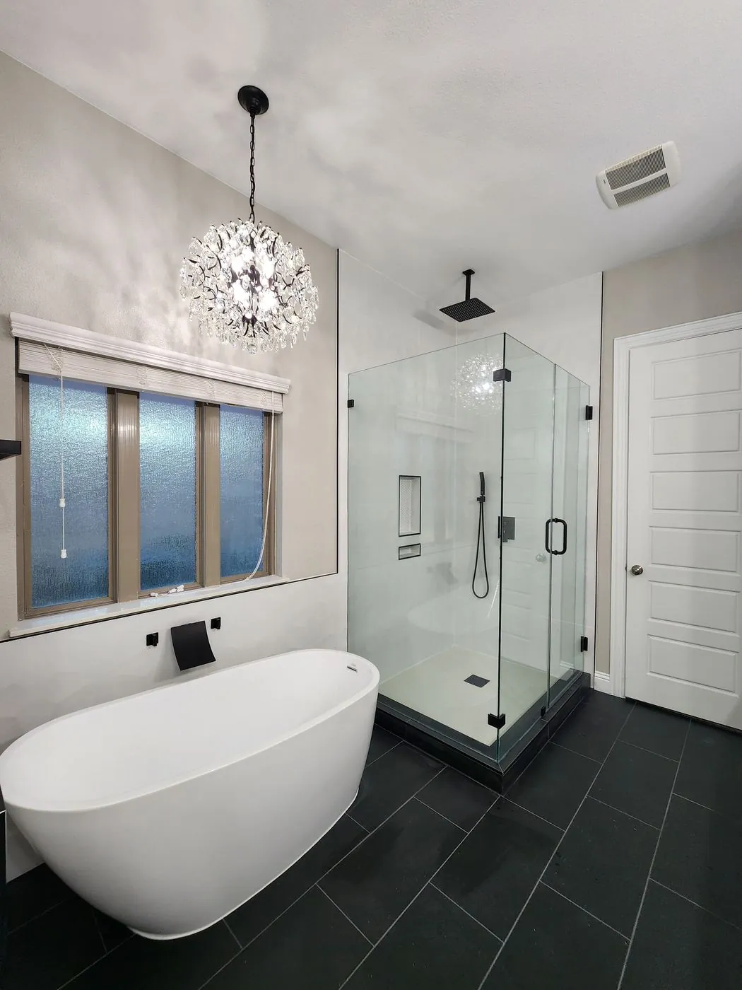 Modern bathroom with a freestanding tub, crystal chandelier and frameless glass shower over dark tile floor