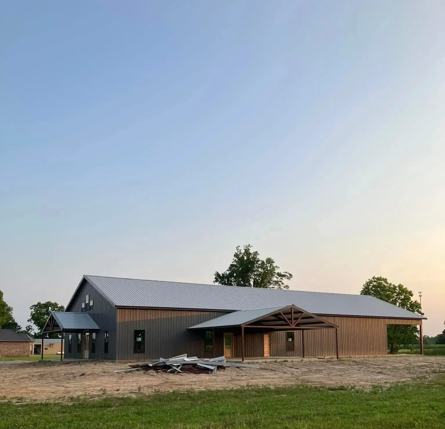Long dark metal barndominium with brown siding, a gabled entry porch and a large truss-style covered porch