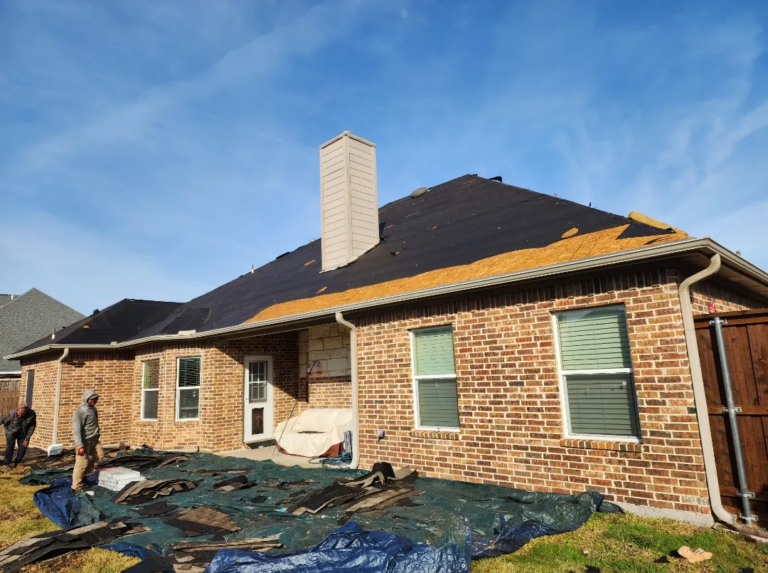 Old shingles removed down to the roof deck with debris collected on tarps behind a brick home